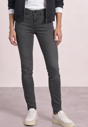 Personne portant un jean skinny gris foncé, des baskets blanches et beiges, une chemise blanche et une veste noire zippée, debout sur un sol gris clair.