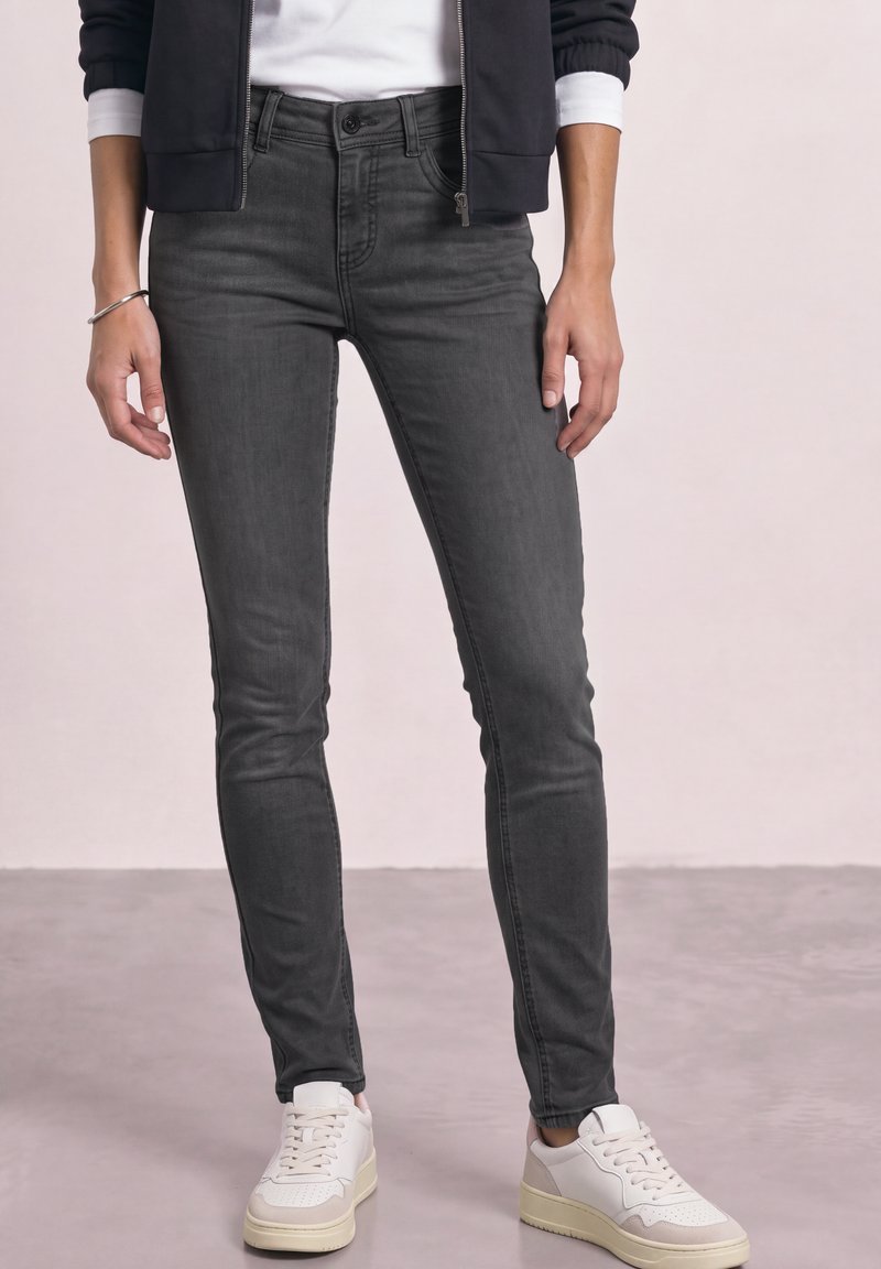 Personne portant un jean skinny gris foncé, des baskets blanches et beiges, une chemise blanche et une veste noire zippée, debout sur un sol gris clair.