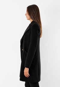 Cappotto in lana nero con vestibilità su misura, caratterizzato da un colletto a revers, tasche laterali e un sottile accento dorato sulla chiusura con bottoni.