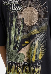 Μαύρο μπλουζάκι με πολύχρωμο γραφικό που απεικονίζει κάκτους, βουνά και ήλιο. Τα σχέδια περιλαμβάνουν πράσινες, κίτρινες και υφασμάτινες λεπτομέρειες.