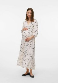 Polka punkta grūtniecības kleita, mīksta auduma, garas piedurknes, dziļš V veida kakla izgriezums, slāņota apakšdaļa, gaismas krēmkrāsas pamatne, melni punkti, kombinēta ar melnām izpildītājām kurpēm.