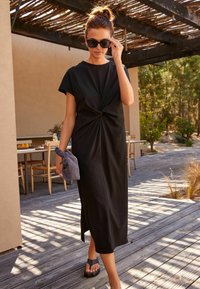 Robe midi noire avec un détail noué à l'avant, manches courtes et une fente sur le côté, associée à des sandales noires et un sac à main gris.