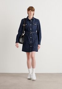 Denim shirt dress in dark blue, featuring long sleeves, button-down closure, belted waist, and two chest pockets. Worn with white sneakers.