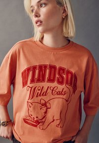 Ülikõrge oranž puuvillane t-särk, millel on tekst "WINDSOR Wild Cats" ja joonistus punase tindiga kausist sööva kassi kohta.