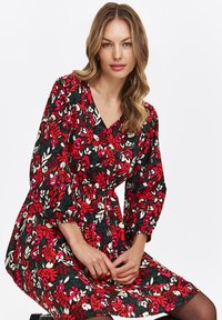 Kobieta z falującymi blond włosami, ubrana w długą, czerwono-czarną i białą sukienkę w kwiaty, siedząca z dłońmi delikatnie złożonymi na kolanach.