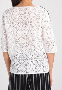 Top in pizzo bianco con intricati motivi floreali, scollatura a giro e maniche a tre quarti, abbinato a pantaloni a righe nere.