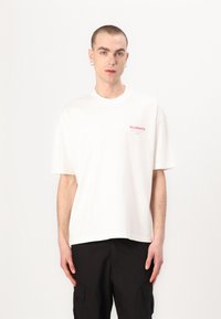 T-shirt en coton blanc à manches courtes, col rond, avec le logo rouge "ALLSAINTS" sur la poitrine gauche. Porté avec un pantalon cargo noir.