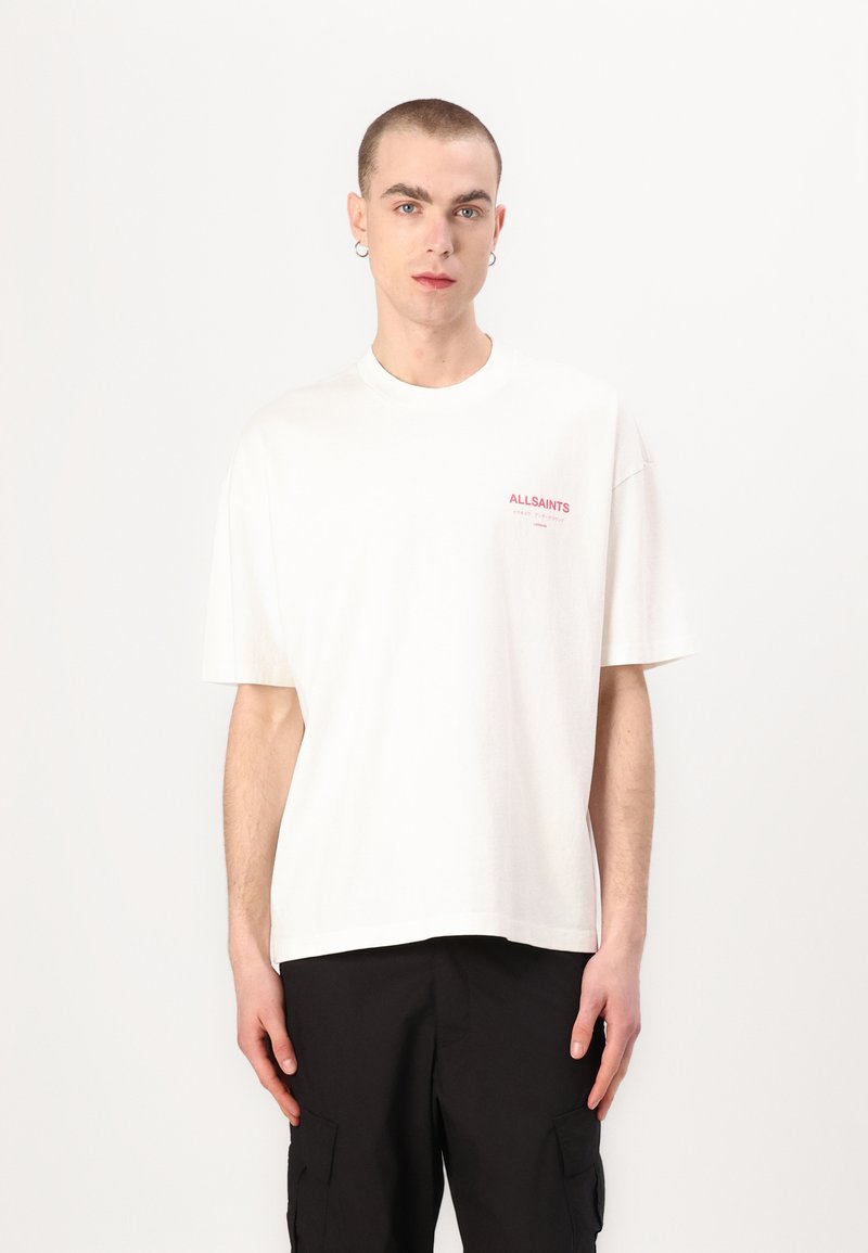 T-shirt en coton blanc à manches courtes, col rond, avec le logo rouge "ALLSAINTS" sur la poitrine gauche. Porté avec un pantalon cargo noir.