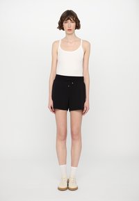 Pantalones cortos negros con cintura elástica y cordón, combinados con una camiseta sin mangas blanca. El modelo se encuentra de pie con calzado neutral y calcetines por el tobillo.