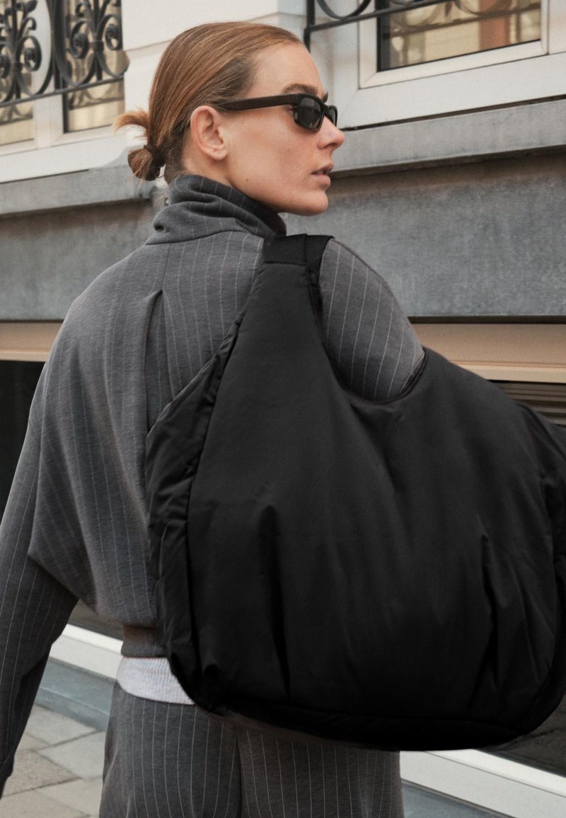 Sac noir à épaulettes rembourrées avec une texture douce et une large sangle, porté sur un ensemble gris à rayures. Design minimaliste, sans logos visibles.