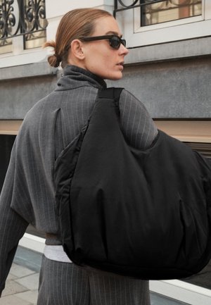 Zwarte puffy schoudertas met een zachte textuur en brede schouderband, gedragen over een grijs gestreept outfit. Minimalistisch ontwerp, geen zichtbare logo's.