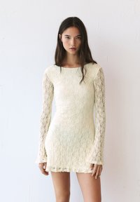 Robe mini en dentelle crème, avec de longues manches en dentelle, un corps ajusté et un ourlet festonné. Motif floral texturé sur l'ensemble.
