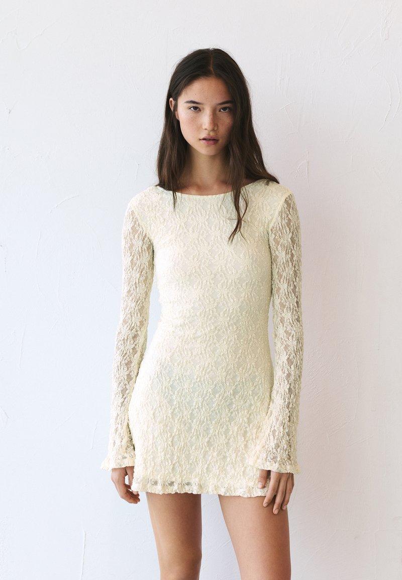 Robe mini en dentelle crème, avec de longues manches en dentelle, un corps ajusté et un ourlet festonné. Motif floral texturé sur l'ensemble.