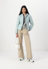 Lyseblå puffer-jakke med glidelåsdesign, elastiske mansjetter, kombinert med beige brede bukser og en grå hettegenser; hvit håndveske og joggesko.