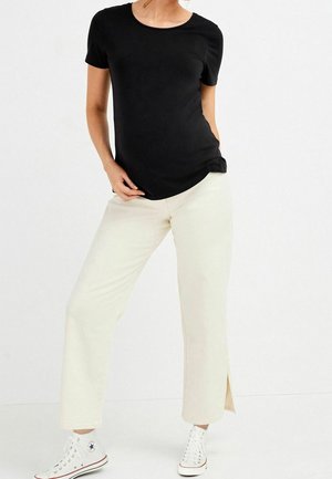 Personne portant un t-shirt noir à manches courtes, un pantalon blanc à jambes larges avec des fentes latérales et des baskets blanches montantes, debout devant un fond uni.