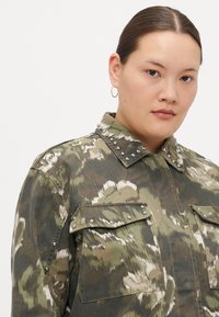 Veste en camouflage avec de grandes poches, des accents cloutés le long du col et des épaules, dans des teintes de vert et de beige avec un tissu texturé.
