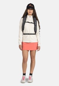 Ljus krämfärgad zip-up jacka, svart ryggsäcksharnäs, klarröd kjol, vita sneakers med rosa detaljer och en svart keps med en bergslogotyp.