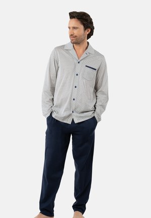 Homme portant une chemise à manches longues à boutons, de couleur claire avec un motif et un col sombre, ainsi qu'un pantalon ample sombre, les mains dans les poches, regardant vers la gauche.