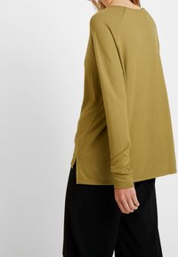 Long-sleeve, olive green top with a rounded neckline, side slits, and soft texture, paired with black pants, shown from the back.