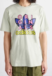 Světle zelené bavlněné tričko s grafickým designem, kde je modrý a černý trojlístek, nápis "ORIGINALS" v růžové a "adidas" v žluté.