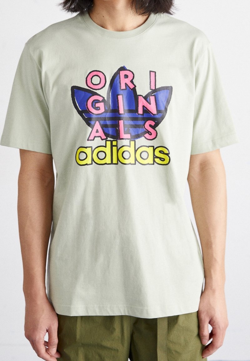 Světle zelené bavlněné tričko s grafickým designem, kde je modrý a černý trojlístek, nápis "ORIGINALS" v růžové a "adidas" v žluté.