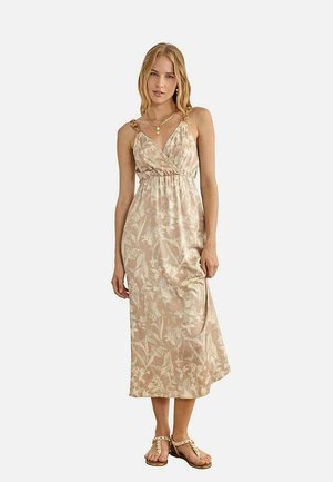 Mujer con vestido midi beige de flores con tirantes finos, collar de cadena dorada y sandalias planas beige, de pie contra un fondo claro.