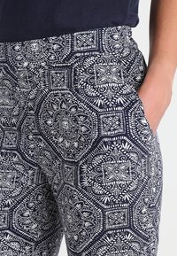 Pantalons extensibles à motif avec fond marine, présentant des designs blancs intriqués, une taille élastique et des poches latérales.