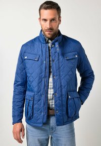 Mand iført en blå quiltet jakke med knappelommer over en ternet skjorte og lyseblå jeans, stående mod en ensfarvet baggrund.