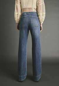 Persoon die een blauw broekje met hoge taille en een doorschijnende crème blouse met kanten details draagt, gezien van achter tegen een grijze achtergrond.