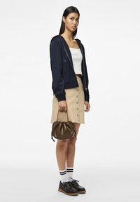 Felpa blu navy sopra una maglietta bianca, gonna beige con bottoni, scarpe nere con suola spessa e una borsa marrone. Il modello sta in piedi con una mano sulla borsa.