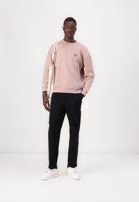 Felpa rosa chiaro con un piccolo logo giallo, abbinata a pantaloni neri e sneakers bianche. Il modello tiene una borsa a tracolla beige.