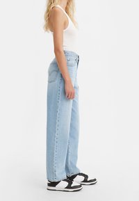 Kvinna står i ljusblå högmidjade vida jeans, vit linne och svart-vita sneakers mot en enfärgad bakgrund.