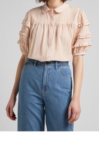 Blusa col collare rosa chiaro in tessuto testurizzato con maniche a volant, abbinata a pantaloni in denim blu a vita alta con vestibilità rilassata.