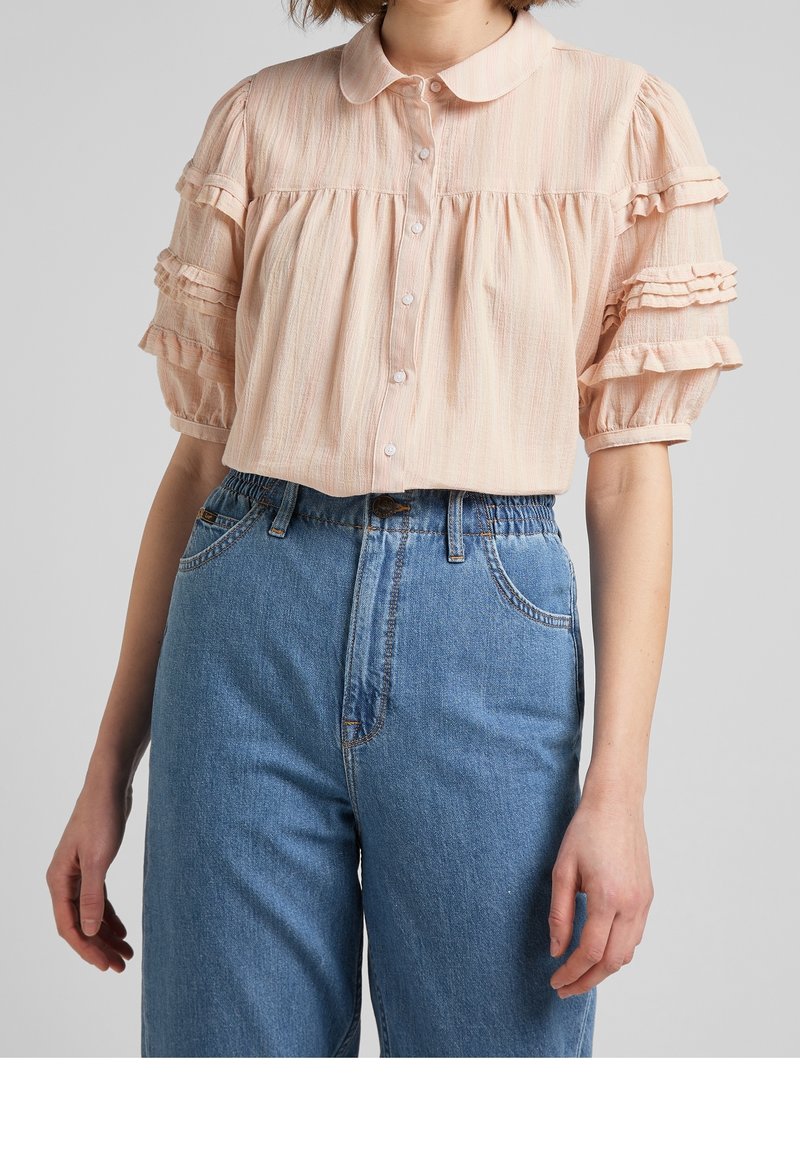 Blusa col collare rosa chiaro in tessuto testurizzato con maniche a volant, abbinata a pantaloni in denim blu a vita alta con vestibilità rilassata.