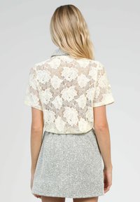 Top en dentelle beige à manches courtes avec un motif floral, superposé sur une jupe grise texturée. La dentelle présente un tissage ouvert et un ourlet en scallop.
