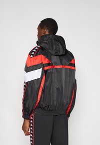 Lekka czarno-czerwona kurtka z kapturem, ozdobiona białymi akcentami i bocznymi paskami. Gładka tekstura z elastycznymi mankietami i dołem.