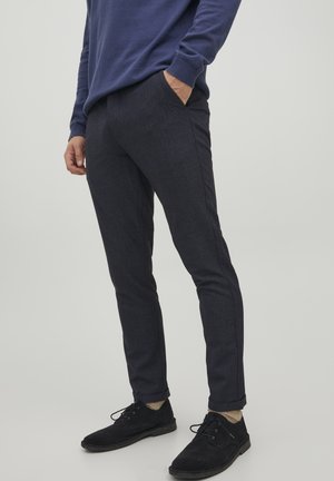 Persoon draagt donkerblauwe slim-fit broek, donkerblauwe trui, beige sokken en zwarte veterschoenen, staat met één hand in de zak.