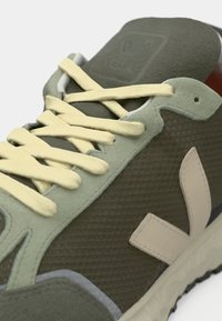 Zaļie režģa sporta apavi ar dzeltenām šņorēm, bēšiem akcentiem un gumijas zoli. Iekļauj teksturētu augšdaļu un logotipu uz mēles.