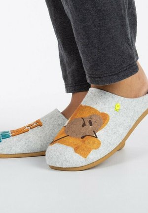 Pies que llevan pantuflas de fieltro grises con personajes de aguacate de dibujos animados, combinados con pantalones gris oscuro sobre un fondo blanco.