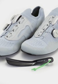 Světle šedé cyklistické boty s pletenou texturou, s nastavením pomocí dials BOA, a černými botníky se zelenou šňůrkou. Podrobný design vzorku podrážky.