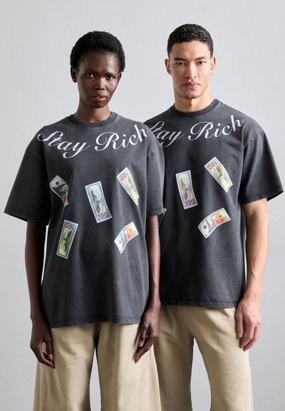 Γκρι υπερμεγέθη T-shirts διαθέτουν λευκό κείμενο "Stay Rich", διακοσμημένα με τυπωμένα χαρτονομίσματα των 100 δολαρίων, κοντά μανίκια και έναν μαλακό βαμβακερό υφή.