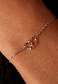 Zilveren en roségouden hartje bedelarmband op een huidkleurige pols, met een delicate ketting en twee verstrengelde hartvormen.