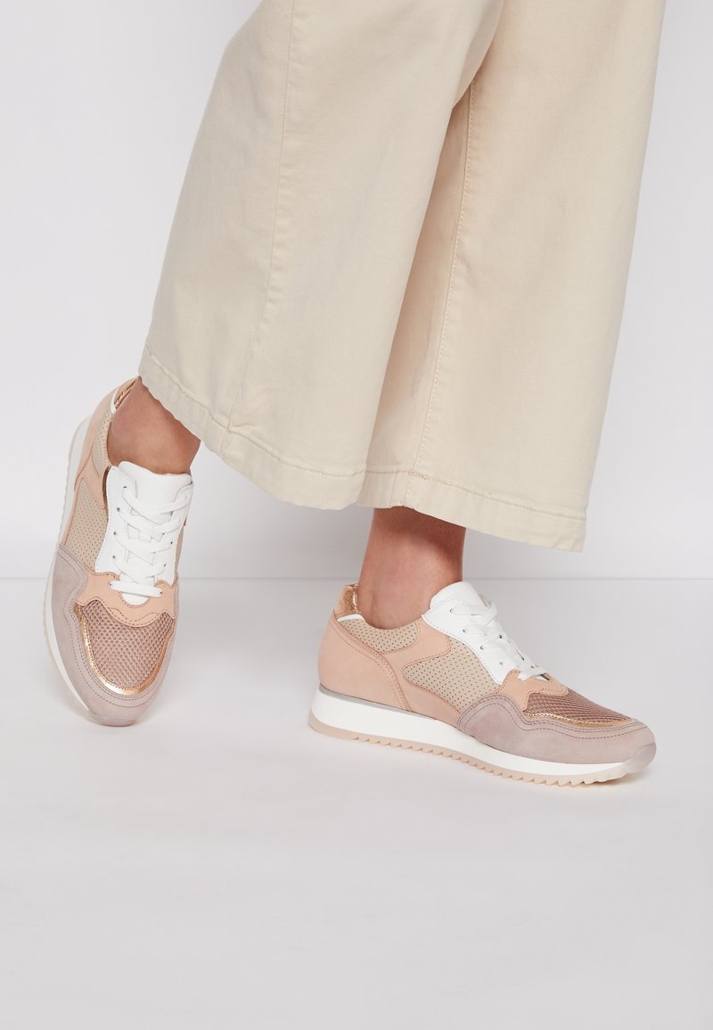 Pieds chaussés de baskets beige et or rose avec des lacets blancs, associés à un pantalon crème à jambes larges sur un fond uni.