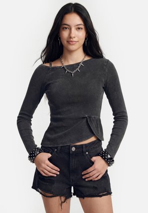 Nuori nainen mustassa pitkähihaisessa paidassa, jossa niitit resoreissa, kuluneissa mustissa farkkushortsissa ja piikillä koristellussa kaulakorussa, seisoo käsineen taskuissa.