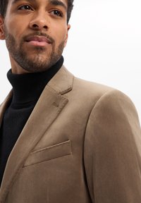 Brązowa zamszowa marynarka z klapami z nacięciem, pojedynczą kieszenią na piersi i poduszkami na ramionach, noszona na czarnym golfie.