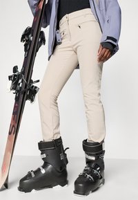 Beige skibroek met een slim fit, voorzien van stikseldetails. Gecombineerd met zwarte sneeuwlaarzen en een snowboard, met een neutrale achtergrond.