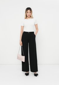 Bijela prilagođena majica, crne široke hlače s remenom i srebrnim detaljima, drži svijetlo ružičasti clutch sa lančanim remenom, crne ravne cipele s mašnom.