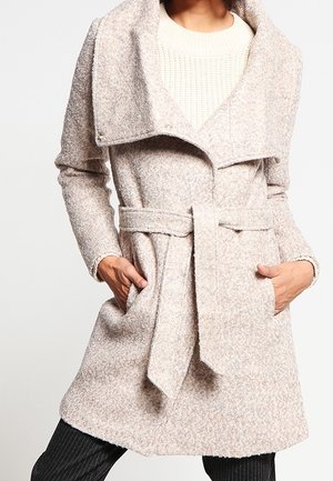 Femme portant un manteau en laine beige ceinturé avec un large col et des manches longues, les mains dans les poches, associé à un pantalon noir à fines rayures.