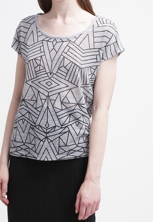 Vrouw draagt een grijs topje met korte mouwen met zwarte geometrische patronen en een zwarte rok, staande tegen een effen witte achtergrond.