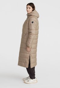 Lång beige puffjacka med huva, med horisontella sömmar, dragkedjor på sidorna och en avslappnad passform. Bärs över svarta byxor och sneakers.